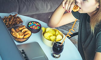 Girl works at a computer and eats fast food. Unhealthy food: chips, crackers, candy, waffles, cola. Junk food, concept.
